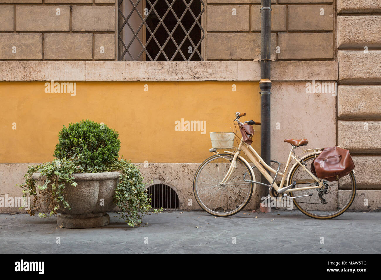 A cream coloured retro style bicycle with brown bags locked up to a drain pipe on a street in Rome, Italy. Stock Photo