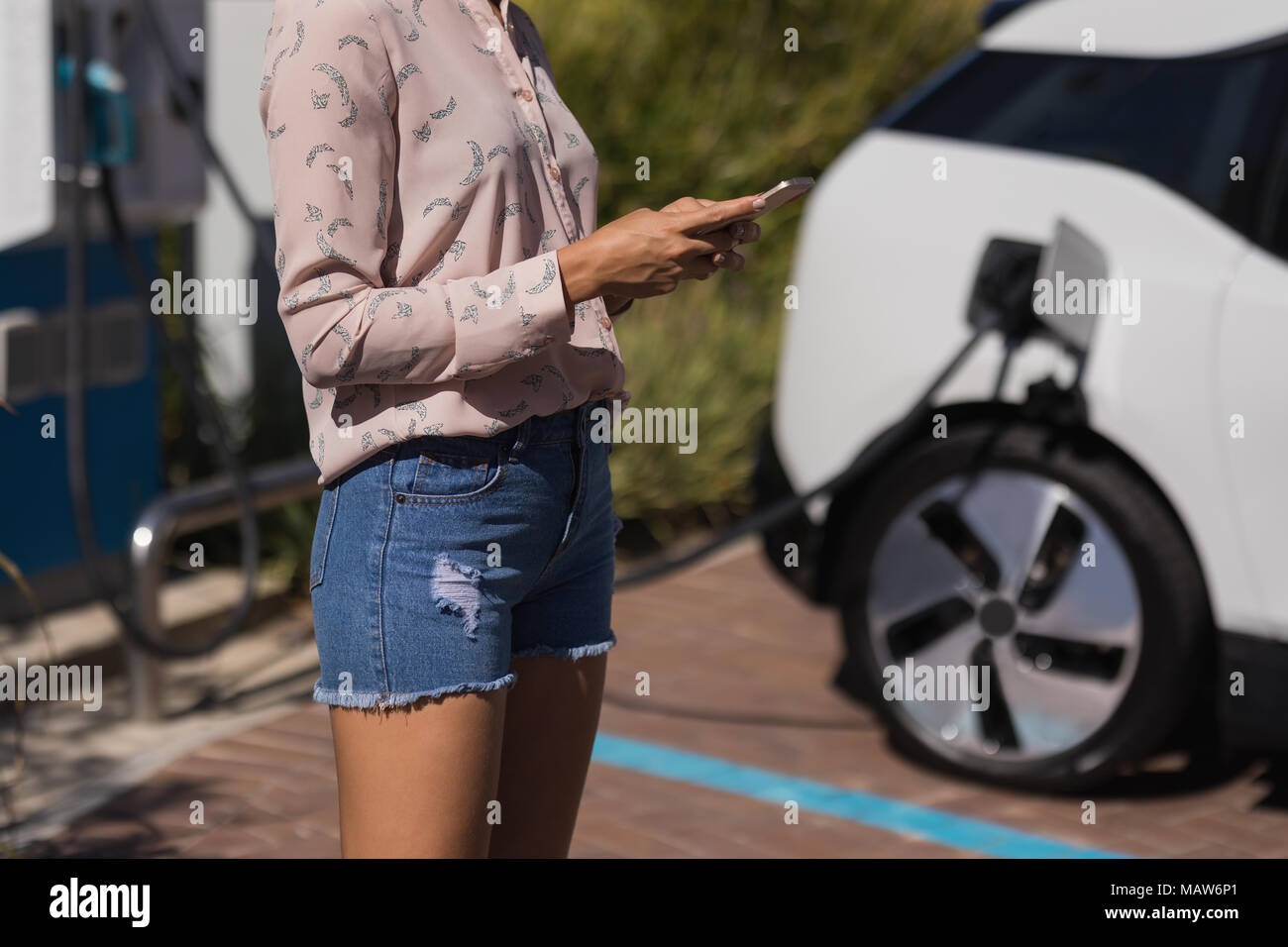 Woman using mobile phone while charging electric car at service station Stock Photo