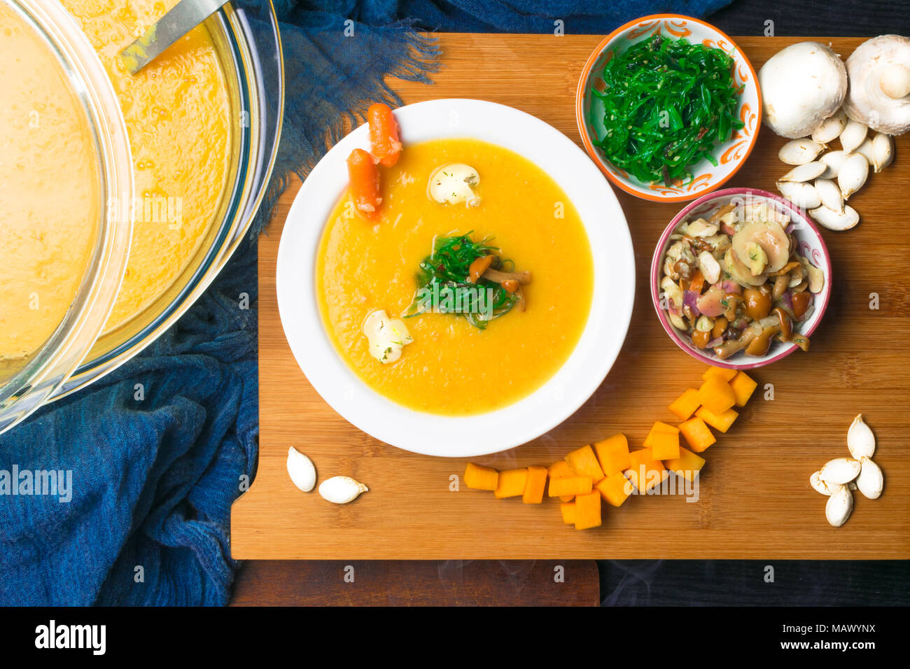 hot pumpkin soup in glass pan and in white plate, decorated with mini carrot, pumpkin seeds, mushroom, honey agaric and green kelp on wooden board. Ve Stock Photo