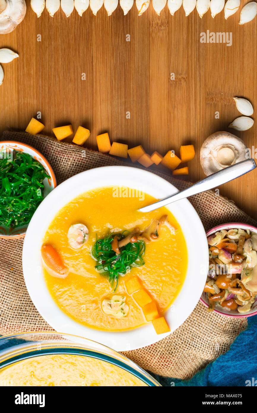 hot pumpkin soup in glass pan and in white plate, decorated with mini carrot, pumpkin seeds line, mushroom, honey agaric and green kelp on wooden boar Stock Photo