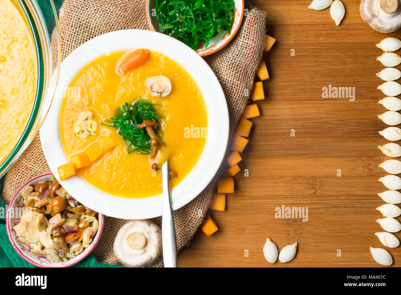 hot pumpkin soup in glass pan and in white plate, decorated with mini carrot, pumpkin seeds line, mushroom, honey agaric and green kelp on wooden boar Stock Photo