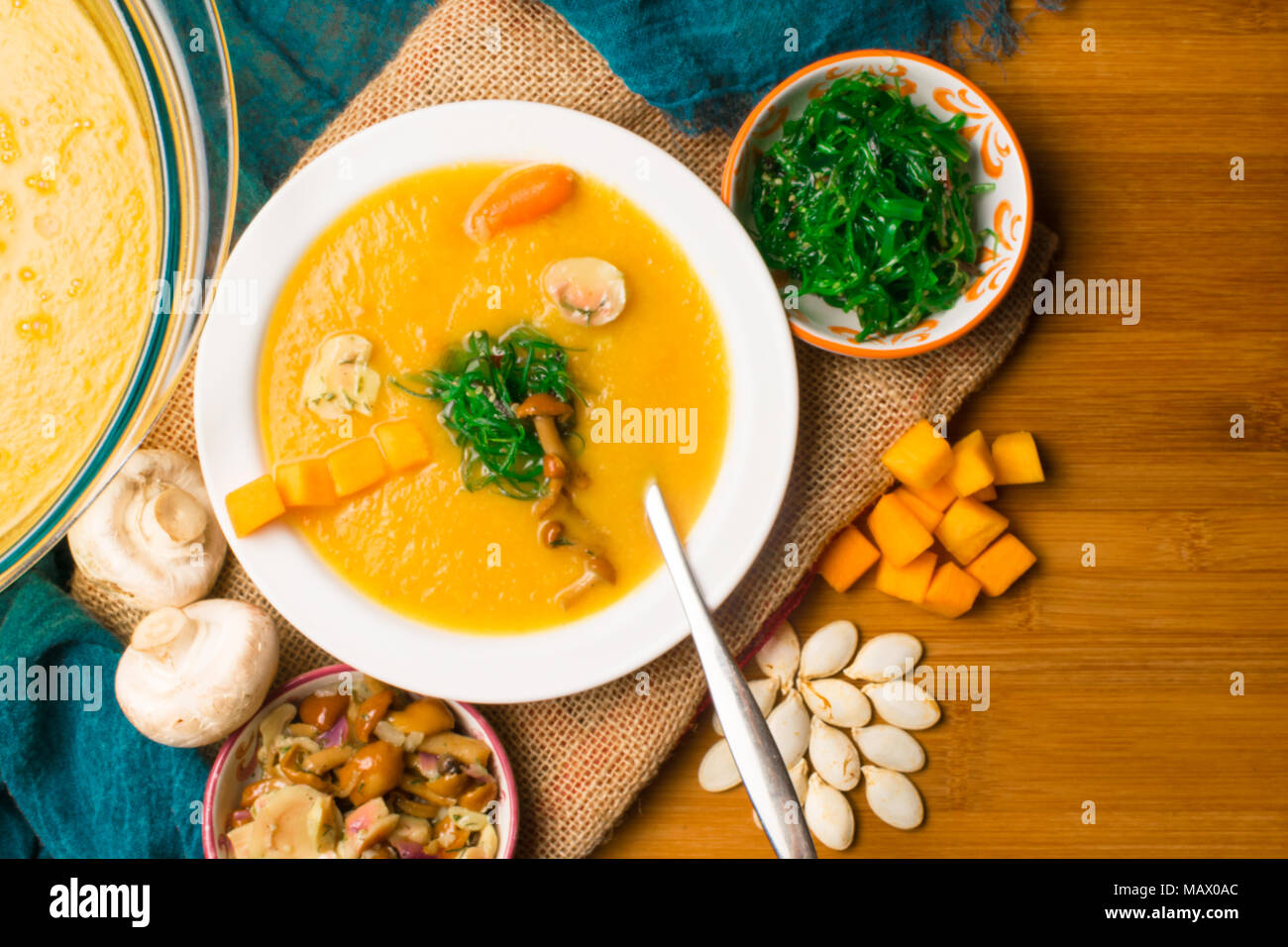 hot pumpkin soup in glass pan and in white plate, decorated with mini carrot, pumpkin seeds, mushroom, honey agaric and green kelp on wooden board. Ve Stock Photo
