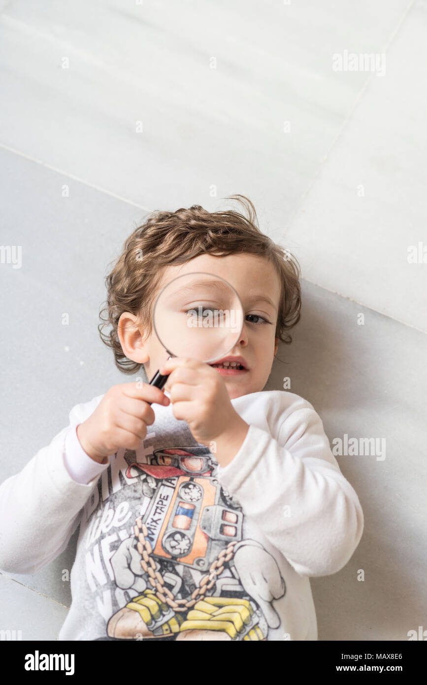 Child lying on a white marble floor looking through a magnifying glass Stock Photo