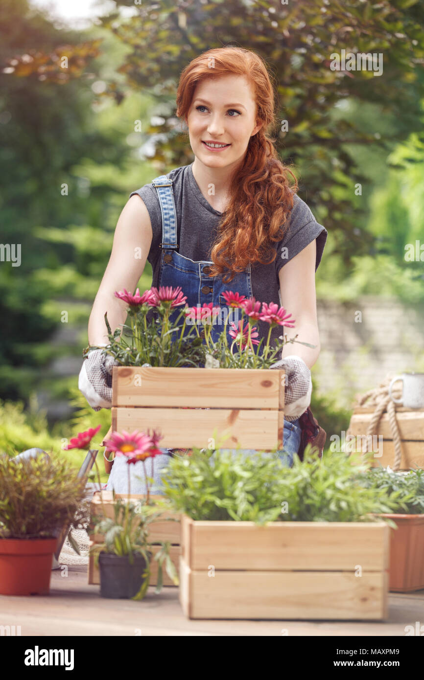 Smiling girl in work trousers, moving a wooden box with flowers while planting in garden Stock Photo