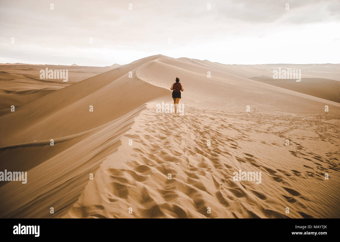Young millennial woman tourist /  traveler walks out across a sand dune into the desert with a red backpack in Huacachina, Peru Stock Photo