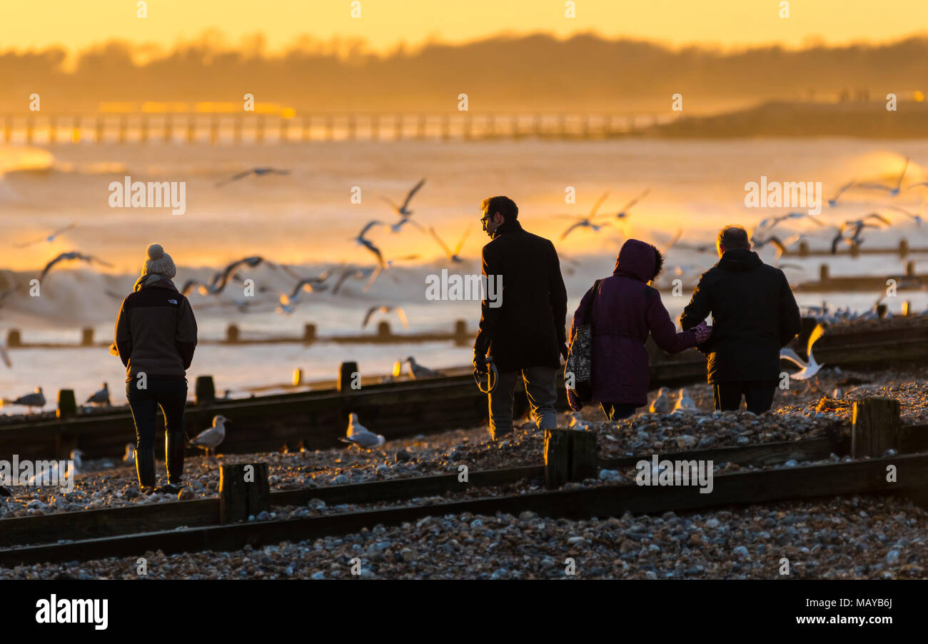 People on a beach at the seaside in the evening as the orange colours of the low sun reflect in the water, in the UK. Stock Photo