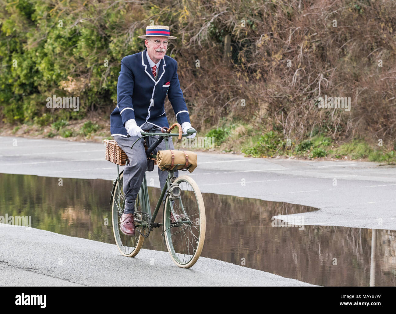 Elderly man cycling on a Victorian bicycle dressed in period costume wearing a blazer and straw boater hat, in the UK. Cyclist riding vintage bike. Stock Photo