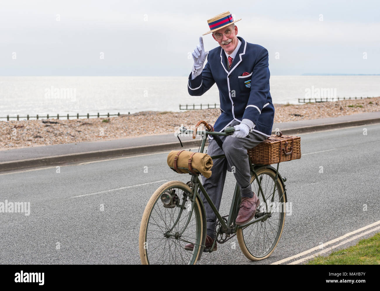 Elderly man cycling on a Victorian bicycle dressed in period costume wearing a blazer and straw boater hat, in the UK. Gentleman tipping hat. Stock Photo