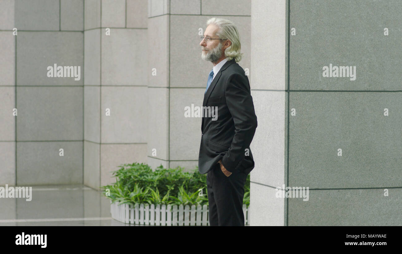 side view of caucasian corporate businessman standing thinking in court yard of modern building, hands in pockets. Stock Photo