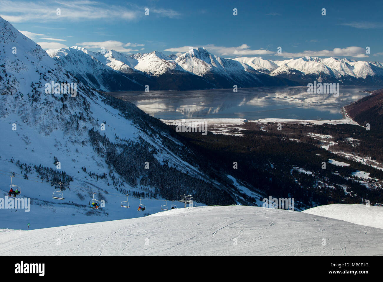 The mountains of the Kenai Peninsula reflect in the calm waters of Turnagain Arm as skiers ride the lift to the top of the run at Alyeska Ski Resort. Stock Photo