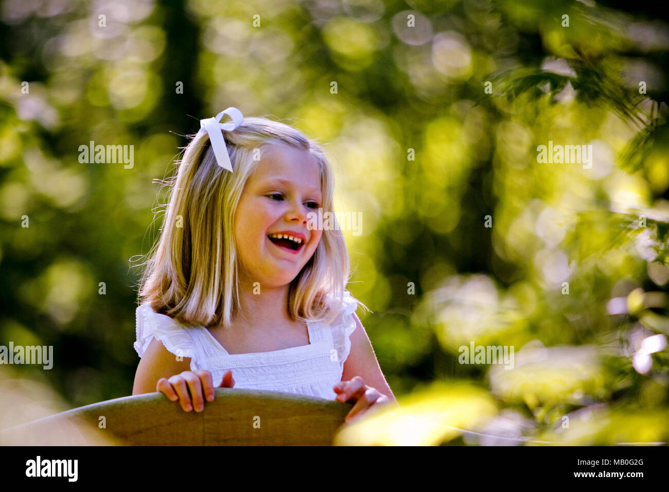 A backlit smiling small Caucasian girl in white dress with blurry green background Stock Photo