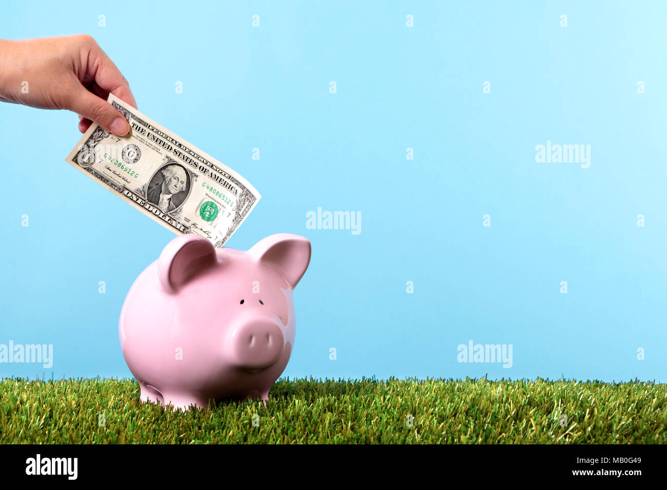 Hand putting a one dollar bill into a pink piggy bank, with grass and blue sky Stock Photo
