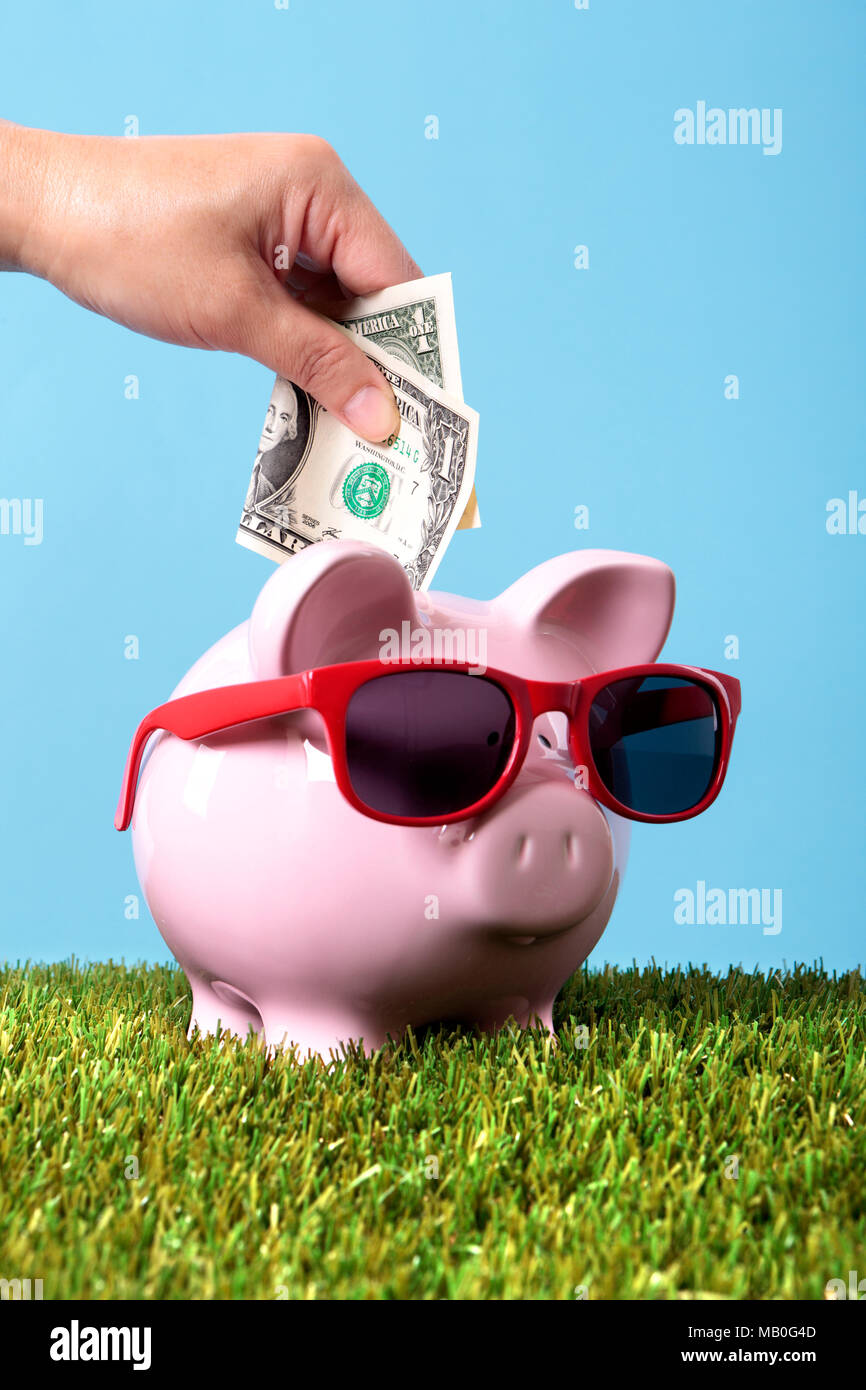 Hand putting a one dollar bill into a pink piggy bank wearing sunglasses. Stock Photo