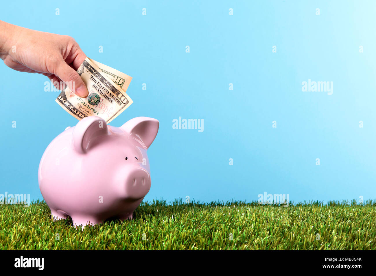 Hand putting a ten dollar bill into a pink piggy bank, with grass and blue sky Stock Photo