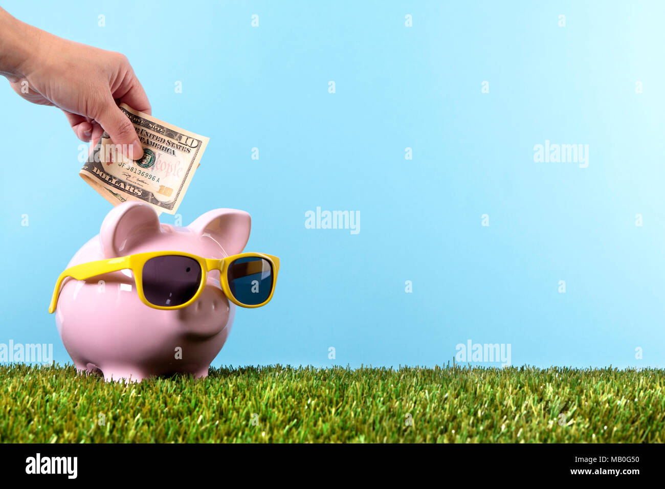 Hand putting a ten dollar bill into a pink piggy bank, with sunglasses, grass and blue sky Stock Photo