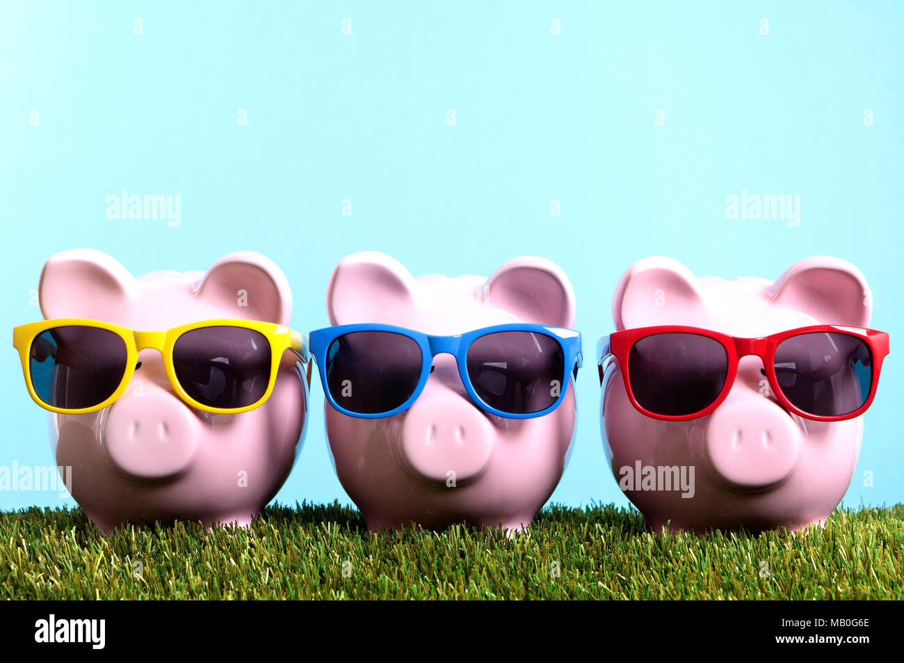 Three pink piggy banks with sunglasses on grass with blue sky.  Studio shot with plain blue background. Stock Photo