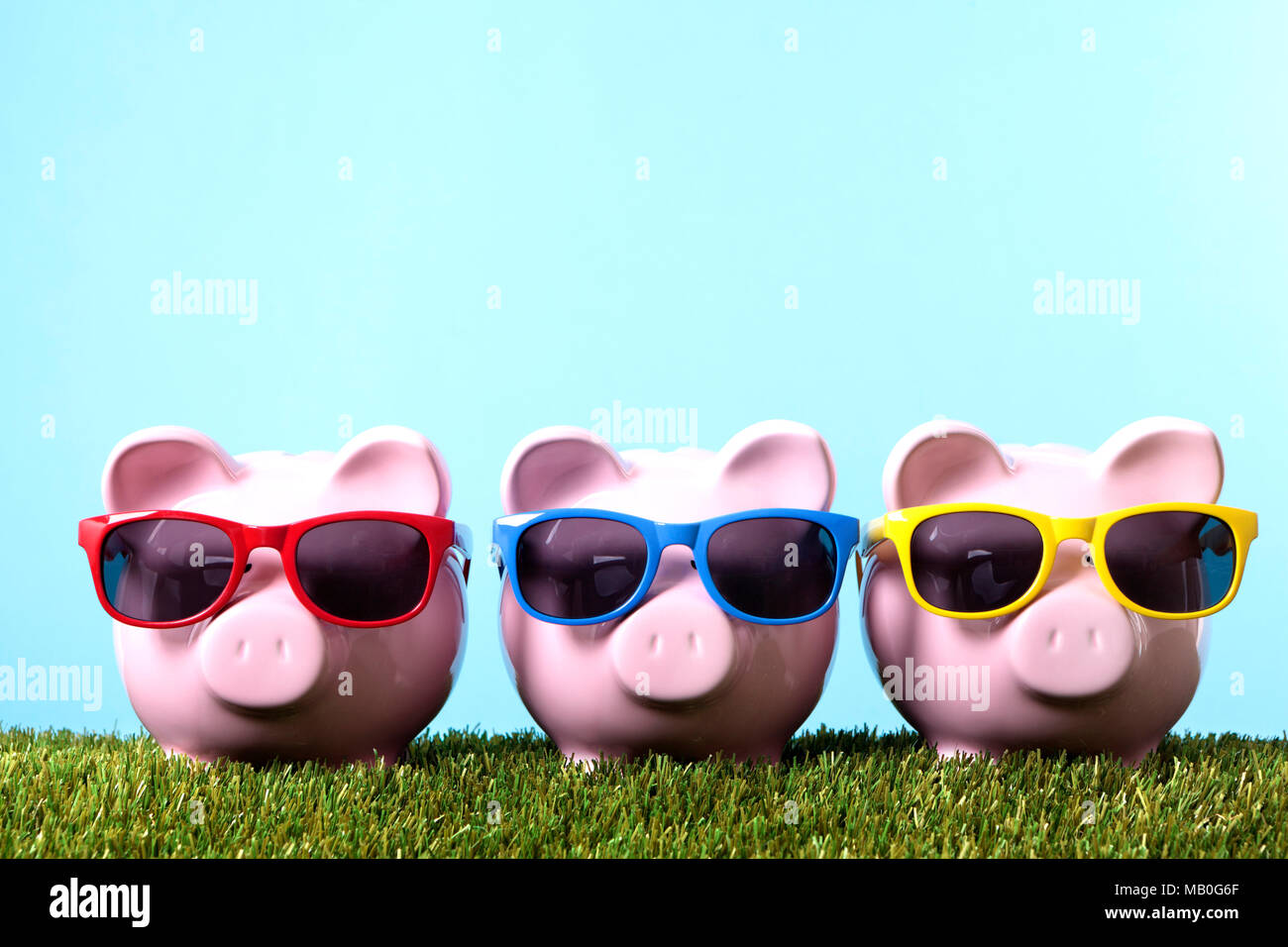 Three pink piggy banks with sunglasses on grass with blue sky.  Studio shot with plain blue background. Stock Photo