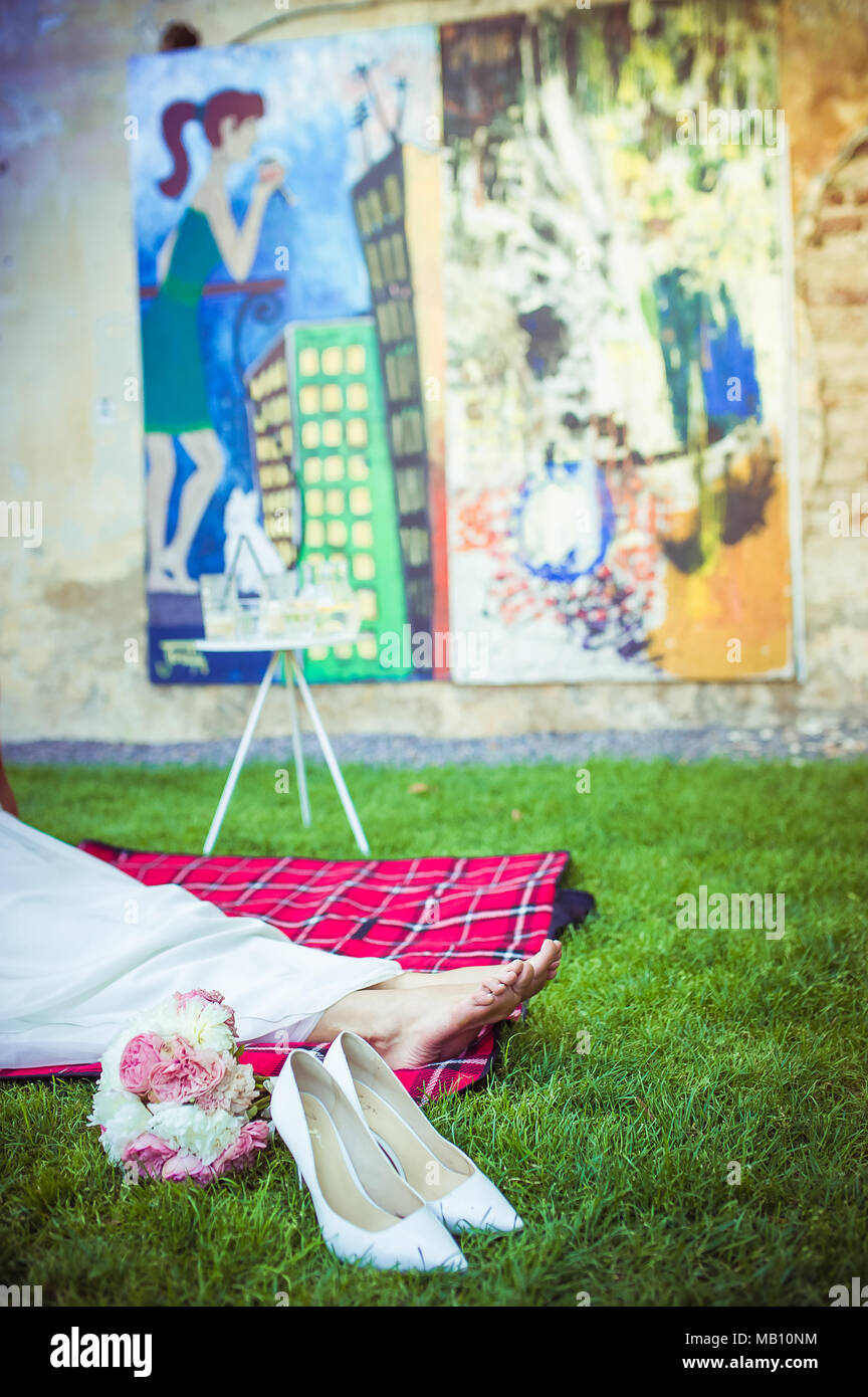 Woman resting her feet on a picnic blanket with colourful paintings in the background. Her shoes and peony bouquet are lying next to her feet Stock Photo