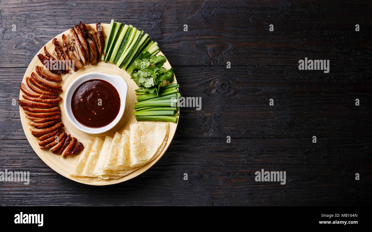 Sliced Peking Duck served with fresh cucumber, green onions, cilantro and roasted wheaten chinese pancakes with Hoysin sauce on black wooden backgroun Stock Photo