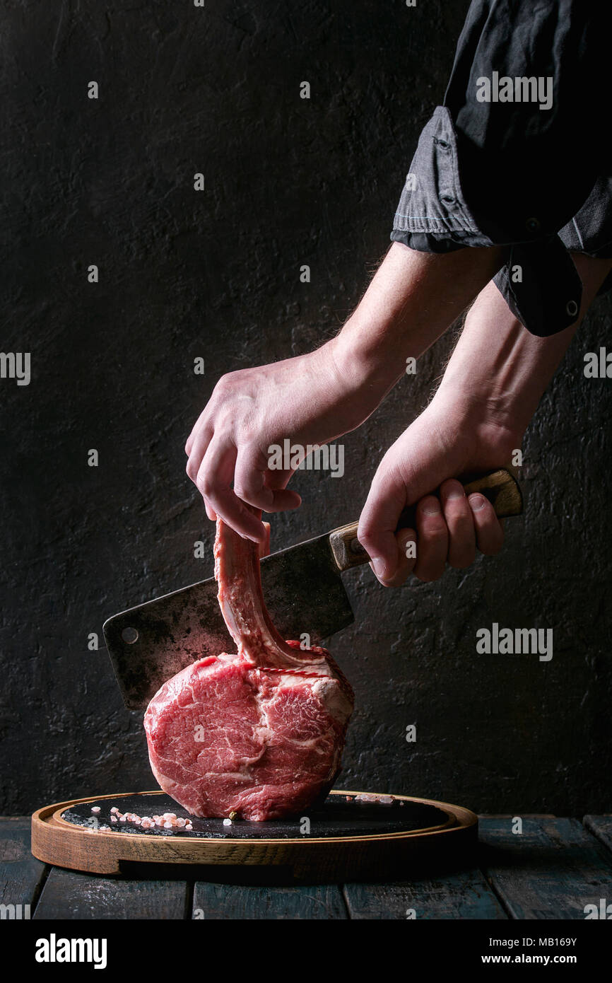 Man's hands cutting raw uncooked black angus beef tomahawk steak on bone by vintage butcher cleaver on round wooden slate cutting board over dark wood Stock Photo