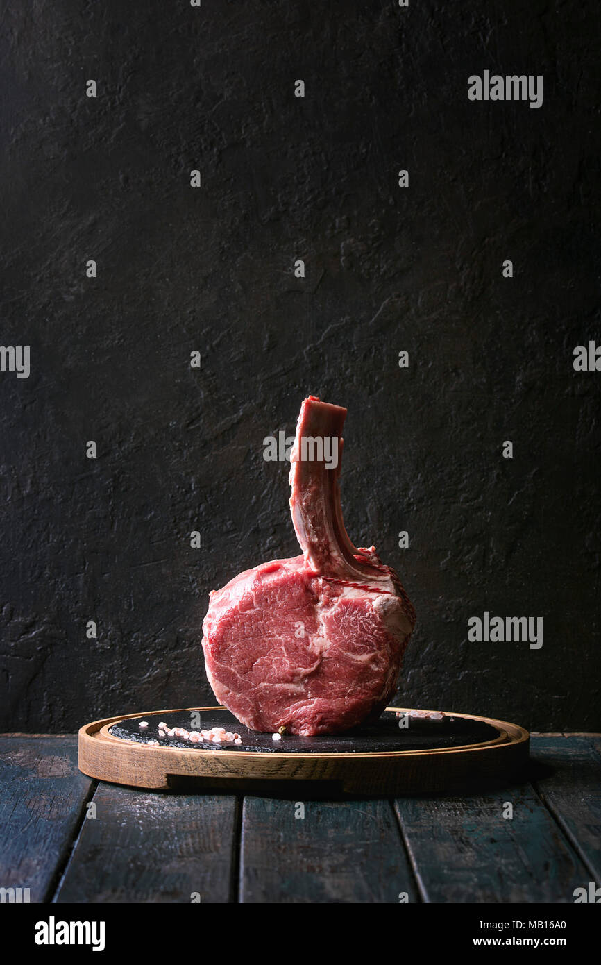 Raw uncooked black angus beef tomahawk steak on bone served with salt and pepper on round wooden slate cutting board over dark wooden plank table. Rus Stock Photo