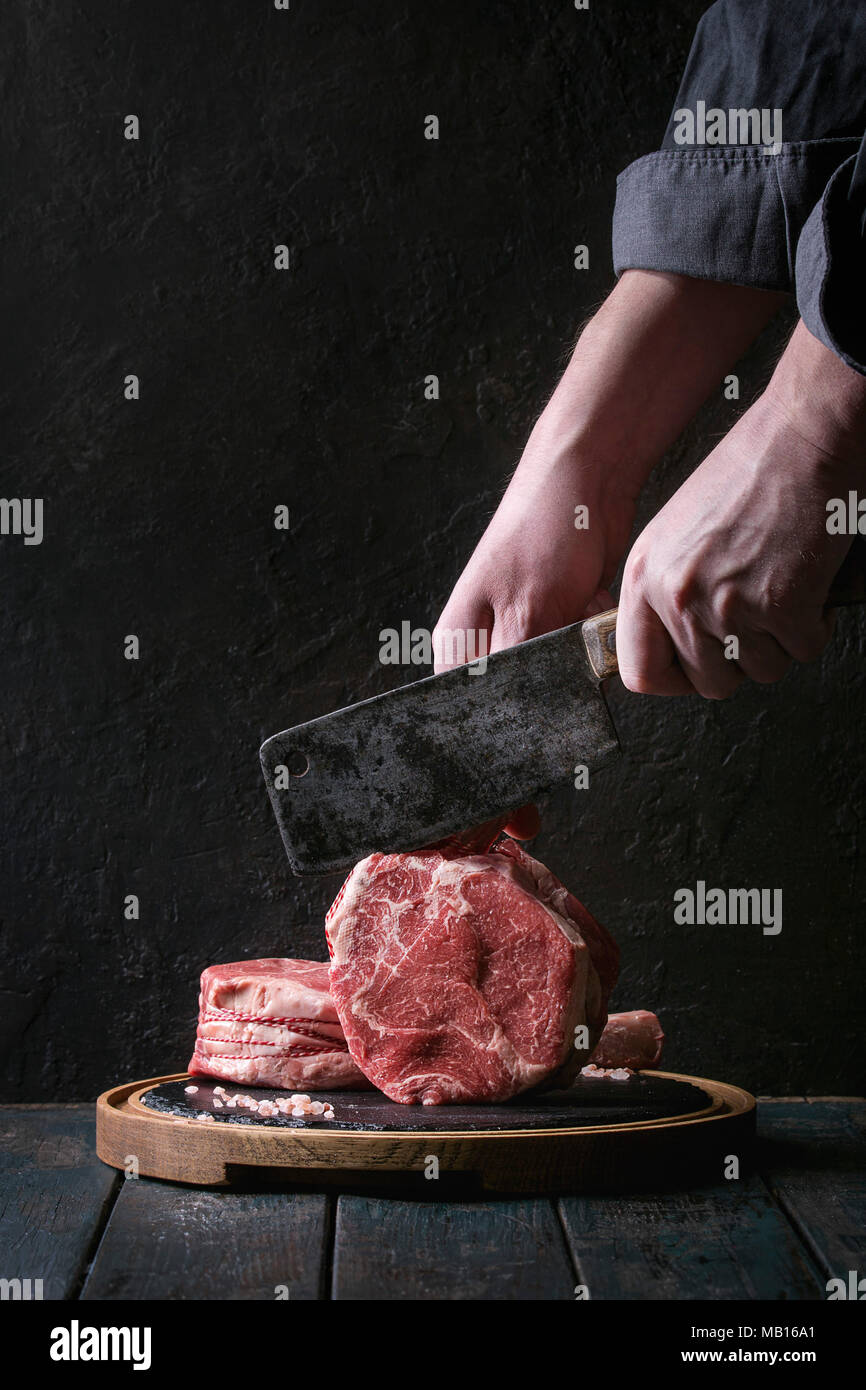 Man's hands cutting raw uncooked black angus beef tomahawk steaks on bones by vintage butcher cleaver on round wooden slate cutting board over dark wo Stock Photo