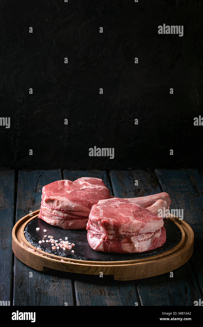 Raw uncooked black angus beef tomahawk steaks on bones served with salt and pepper on round wooden slate cutting board over dark wooden plank table. R Stock Photo