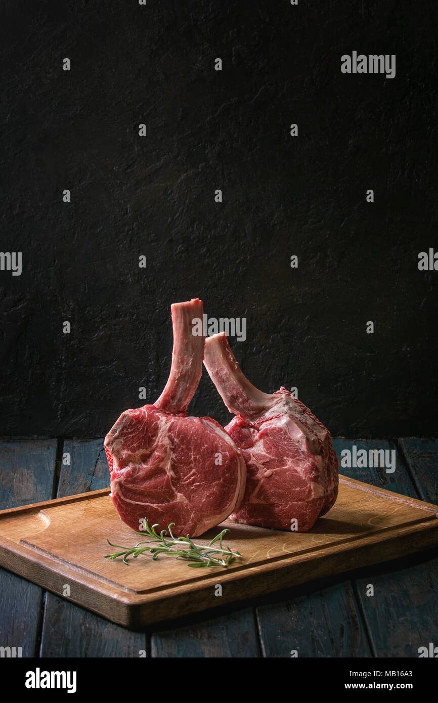 Raw uncooked black angus beef tomahawk steaks on bones served with rosemary, salt and pepper on wooden cutting board over dark wooden plank table. Rus Stock Photo