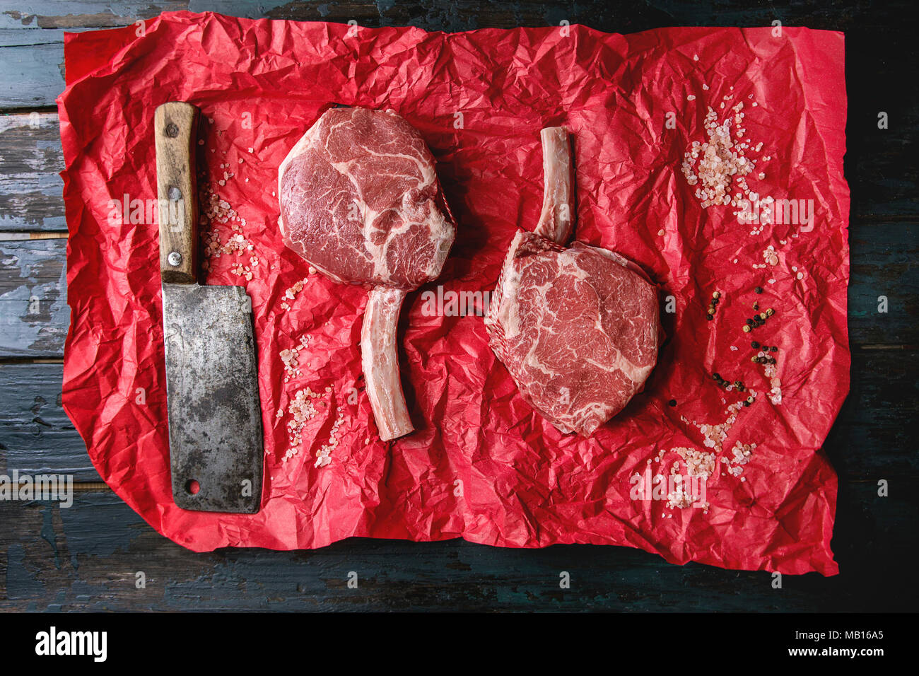 Raw uncooked black angus beef tomahawk steaks on bones served with salt pepper, vintage butcher cleaver on red crumpled paper over dark wooden plank b Stock Photo