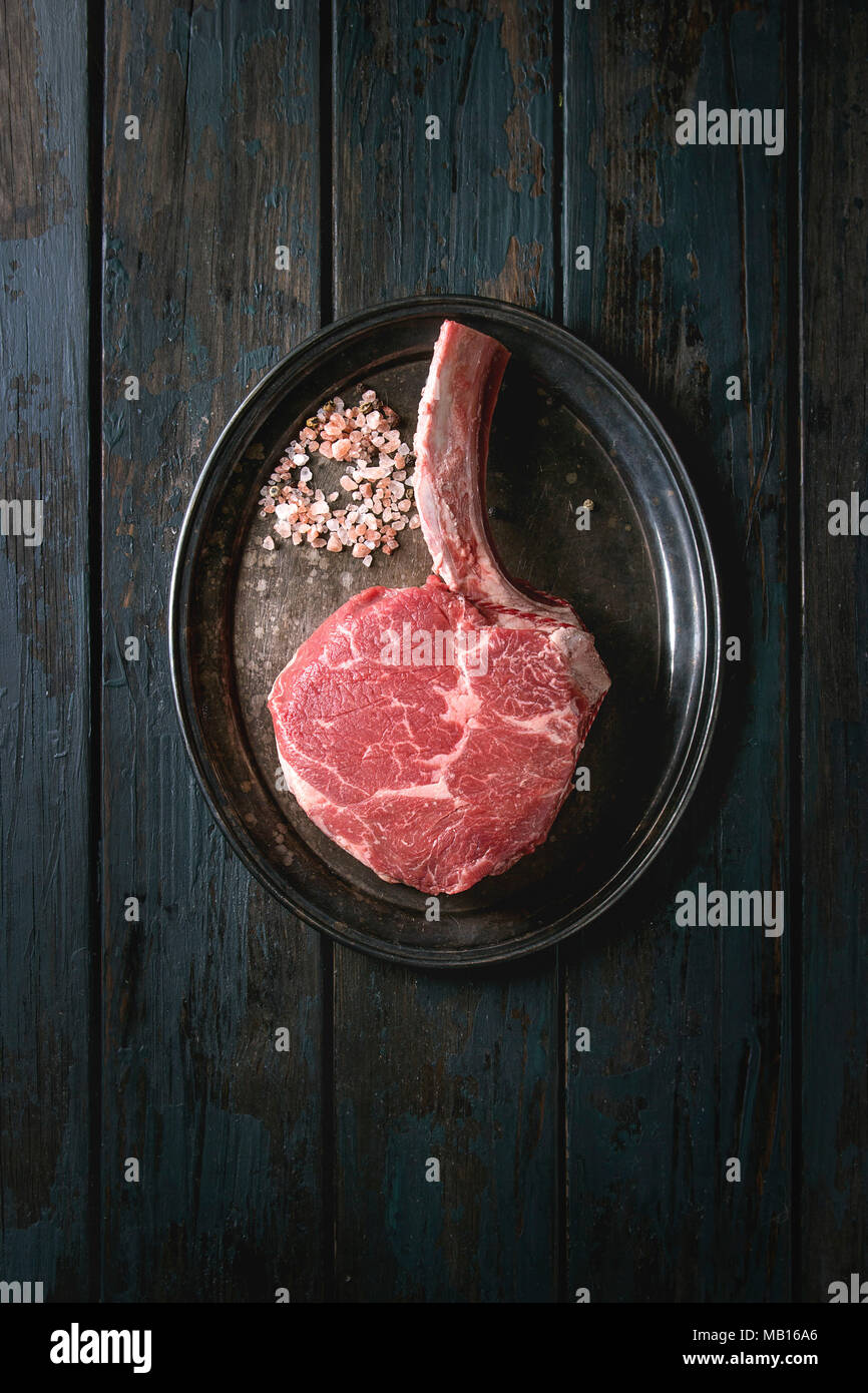 Raw uncooked black angus beef tomahawk steak on bone served with salt and pepper on vintage metal tray over dark wooden plank background. Top view, co Stock Photo