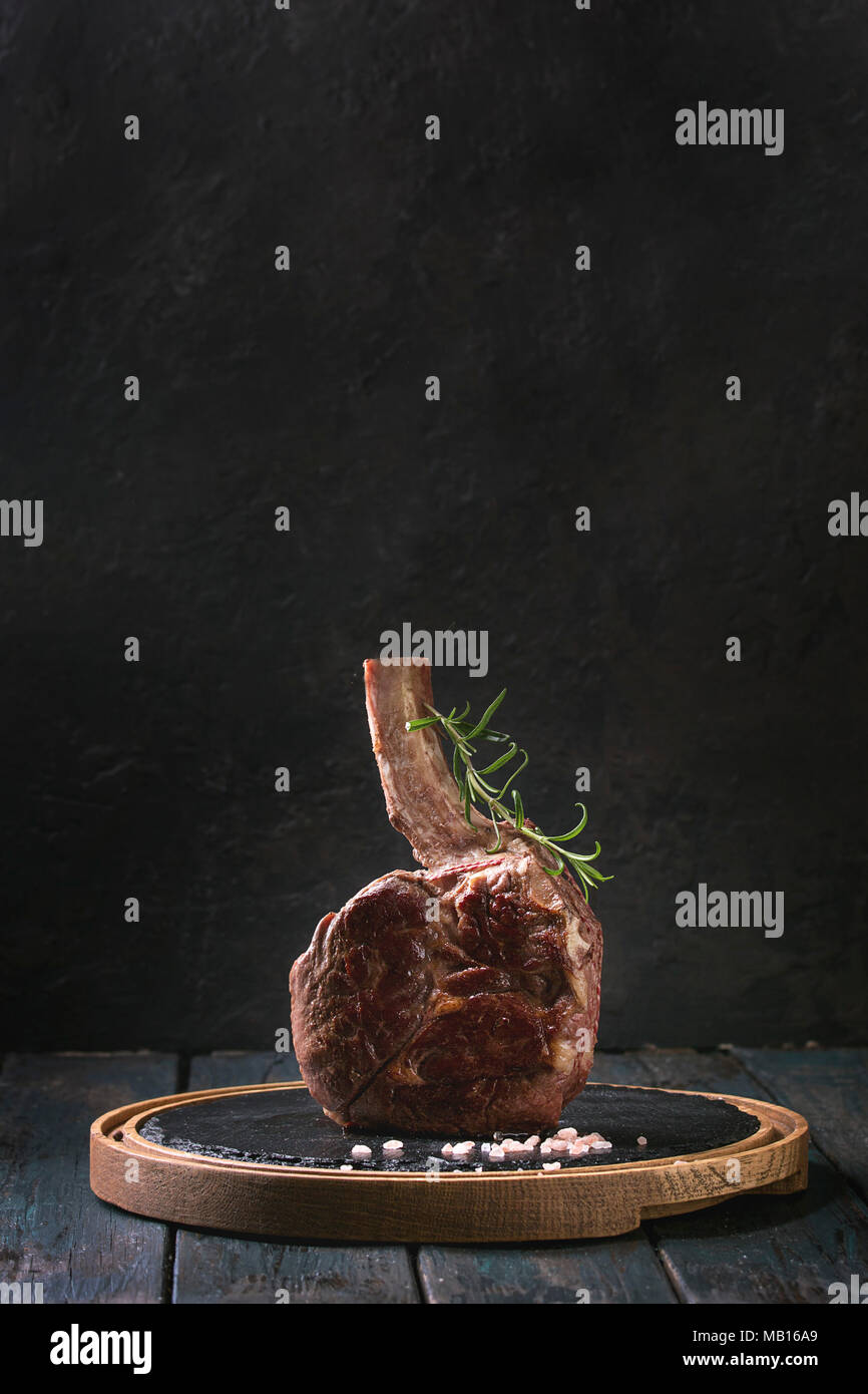 Grilled black angus beef tomahawk steak on bone served with salt, pepper and rosemary on round slate cutting board over dark wooden plank kitchen tabl Stock Photo