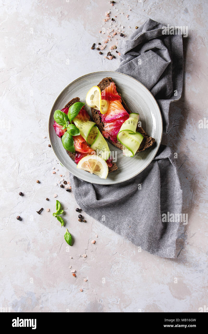 Sliced beetroot marinated salmon sandwiches with rye bread, cucumber, basil and lemon served on ceramic plate with textile napkin over grey texture ba Stock Photo