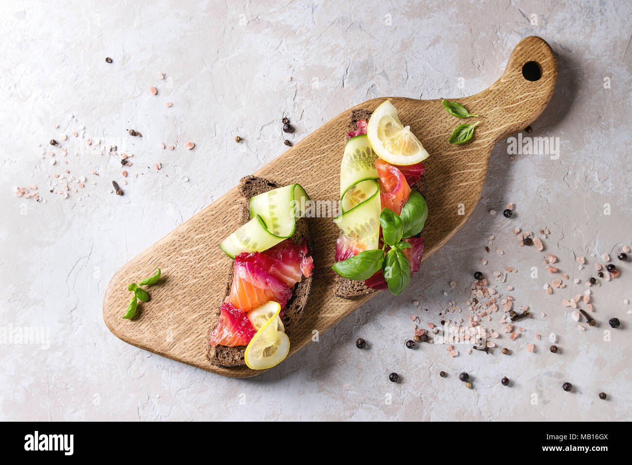 Sliced beetroot marinated salmon sandwiches with rye bread, cucumber, basil and lemon served on wooden cutting board over grey texture background. Top Stock Photo