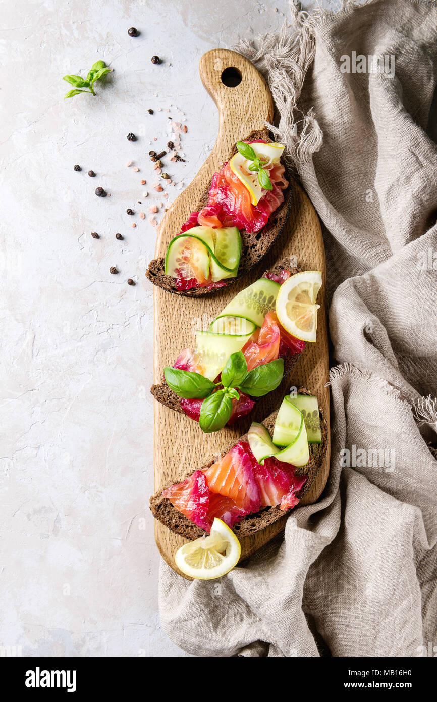 Sliced beetroot marinated salmon sandwiches with rye bread, cucumber, basil and lemon served on wooden cutting board over grey texture background with Stock Photo