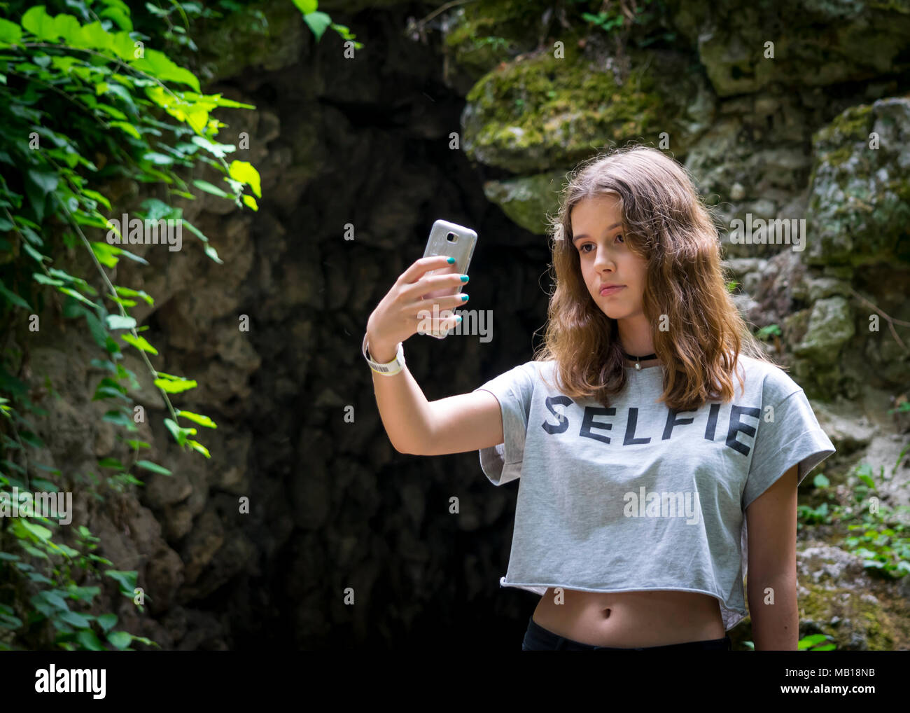 Pretty, teen girl portrait, long hair, outdoors, takes selfie with phone. Portrait of beautiful Caucasian teenage girl in grey top with selfie sign. Stock Photo