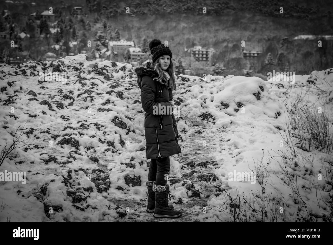 Pretty teen girl in snow, facing camera, black and white. Beautiful Caucasian teenage girl in black coat, snowy mountains in the background. Stock Photo