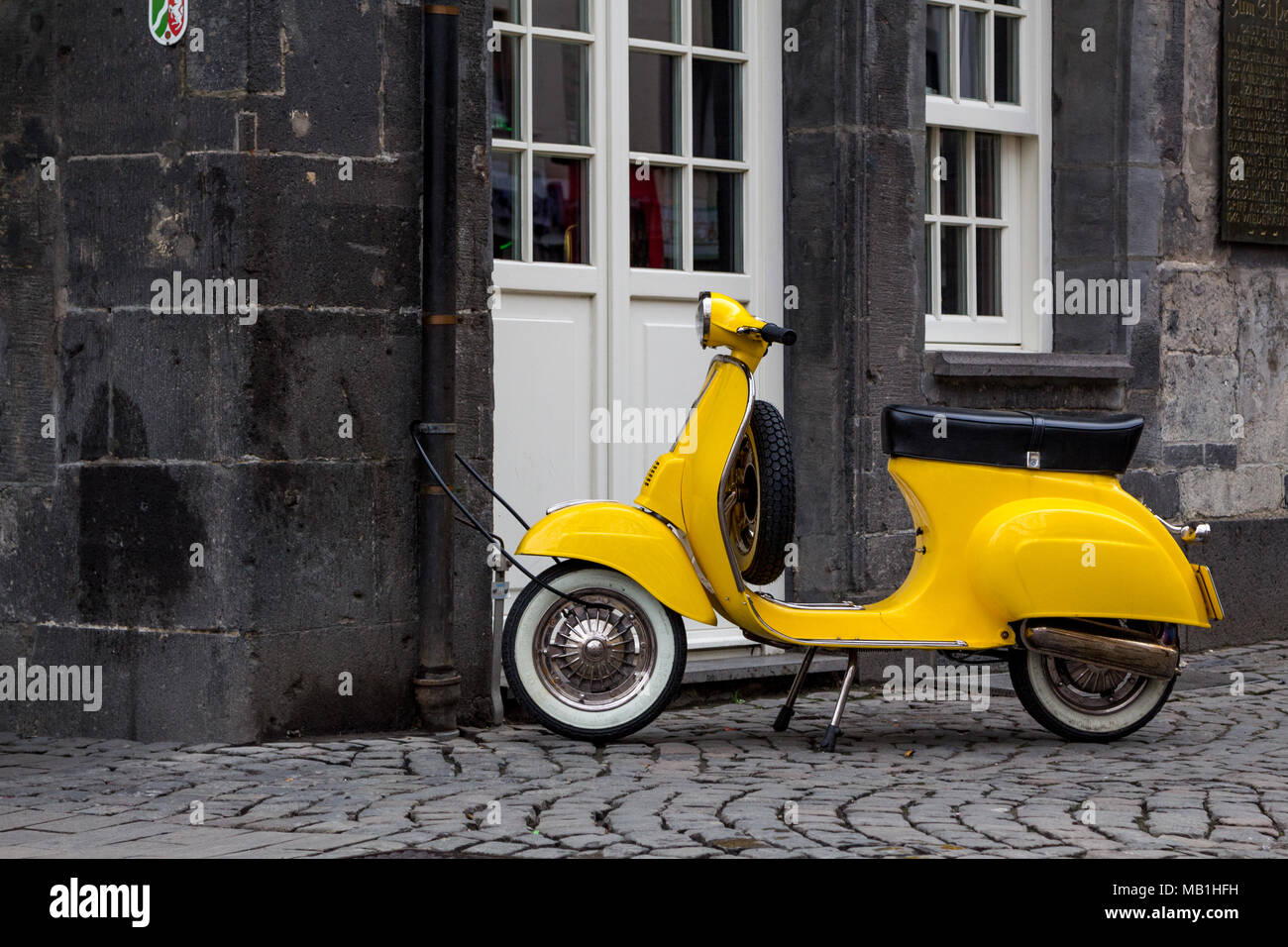 A yellow Italian retro style scooter locked to a rain pipe  of a historic house in the center of the german city of Essen. Stock Photo