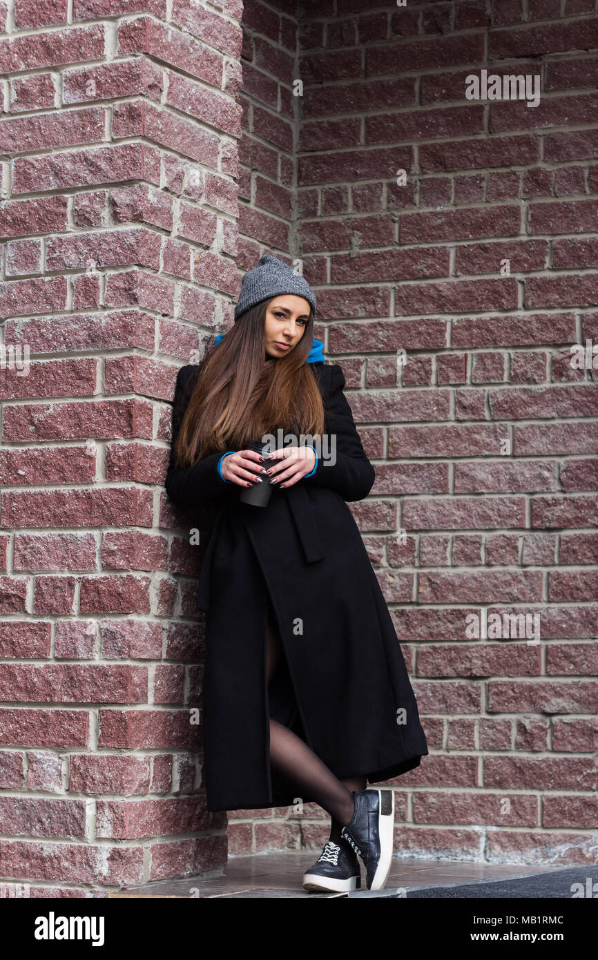stylish young woman weared long black coat and gray knitted hat with coffe to go against brick wall Stock Photo