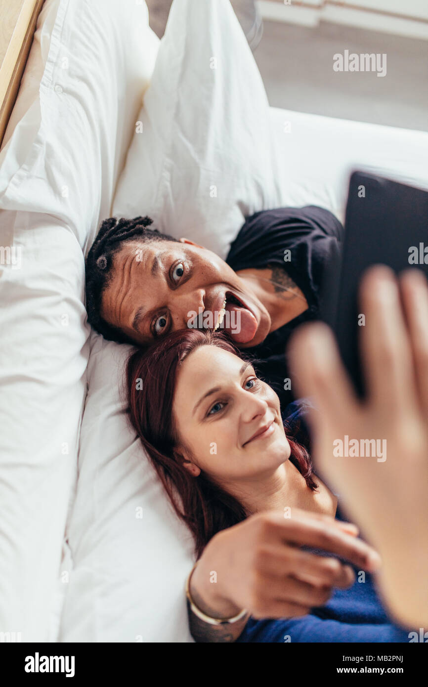 Top view of multiracial couple lying together on bed and taking selfie. Man sticking out tongue with woman taking selfie using mobile phone. Stock Photo
