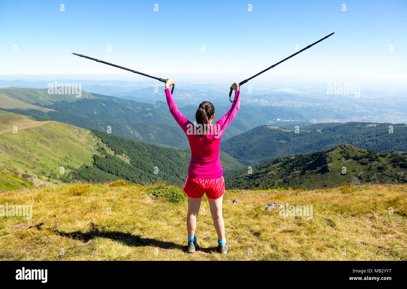 Young woman in pink trekking in the mountain. Enjoying the freedom nature gives. Sunny warm summer day. Holding walking canes in the air. From her bac Stock Photo