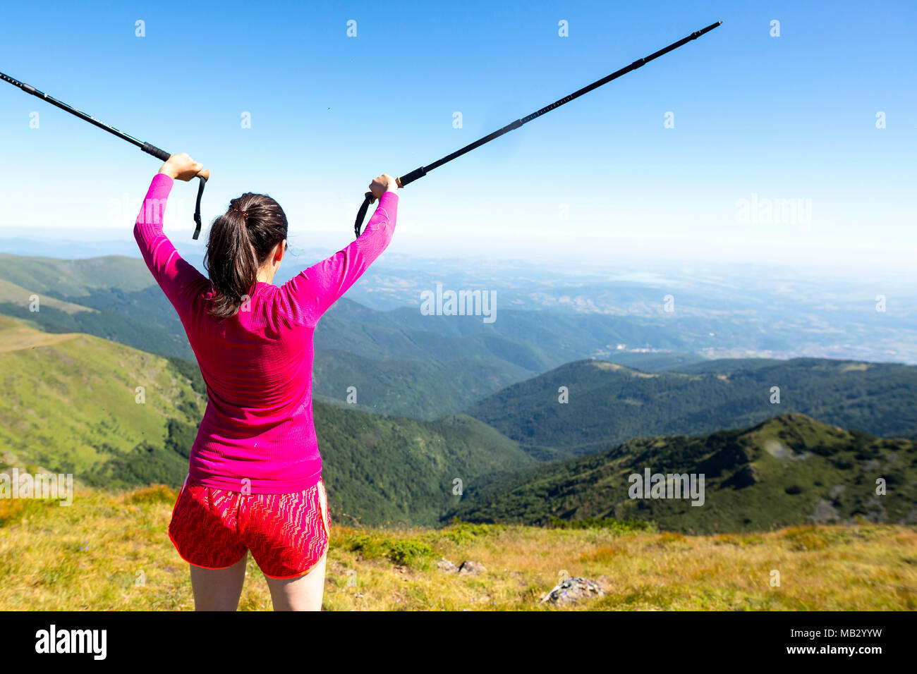Young woman in pink trekking in the mountain. Enjoying the freedom nature gives. Sunny warm summer day. Holding walking canes in the air. From her bac Stock Photo