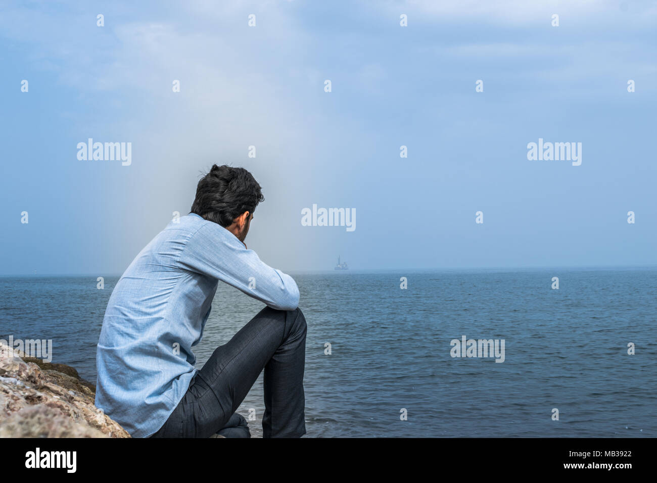 a lonely depressed man after divorced is sitting on the sea rocks and looking to the sea wearing shirt and black paint Stock Photo