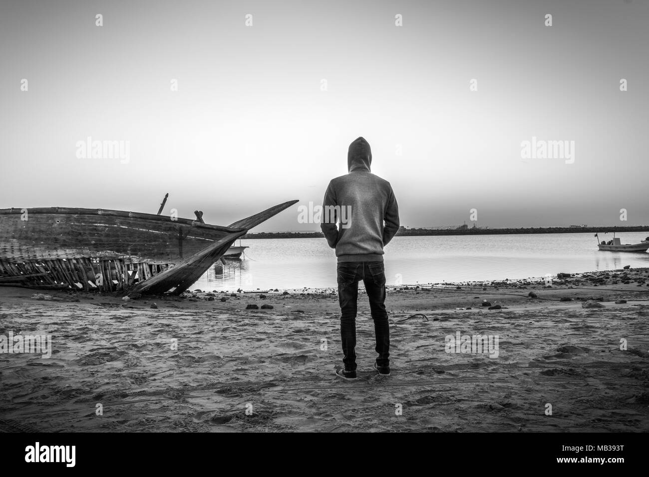 a man wearing hoodie is standing at a beach front of an abandoned broken wooden fishing boat on the side of the beach and looking so sad Stock Photo