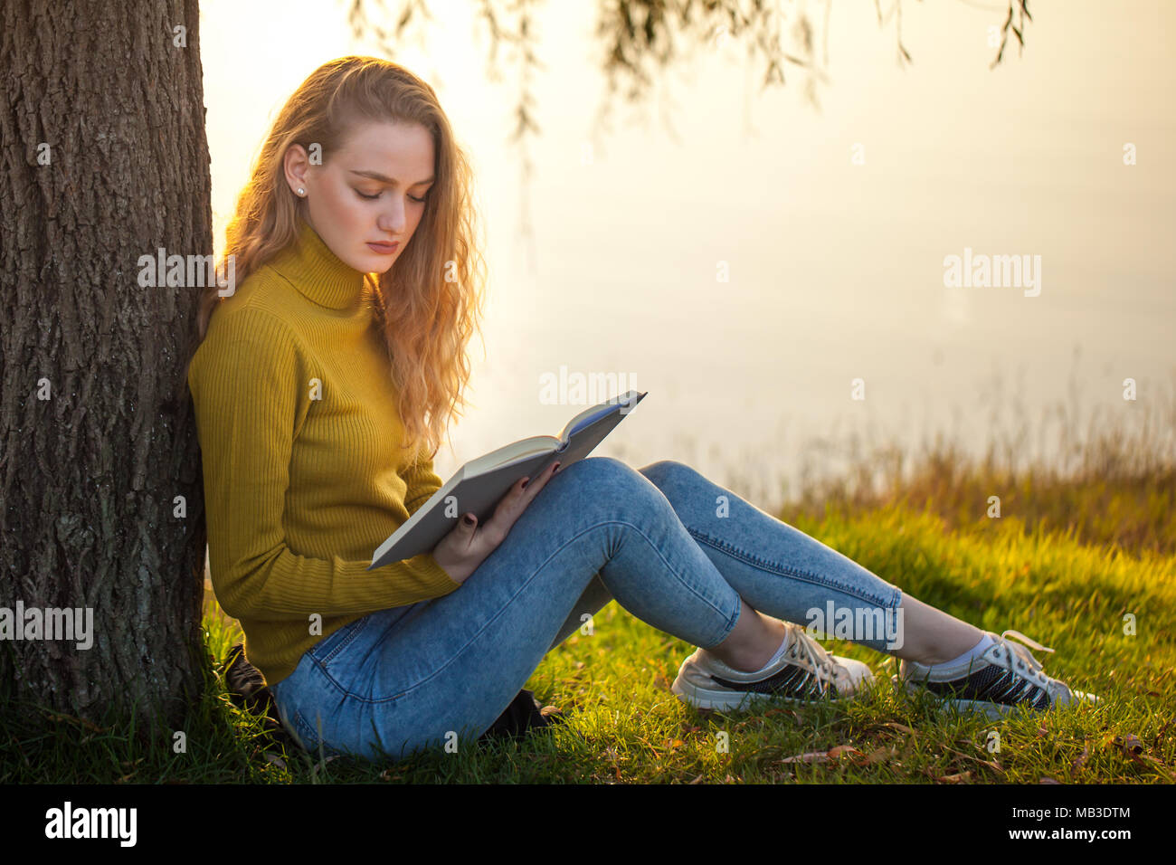 Beautiful young blonde woman sitting and reading book in a park near ...