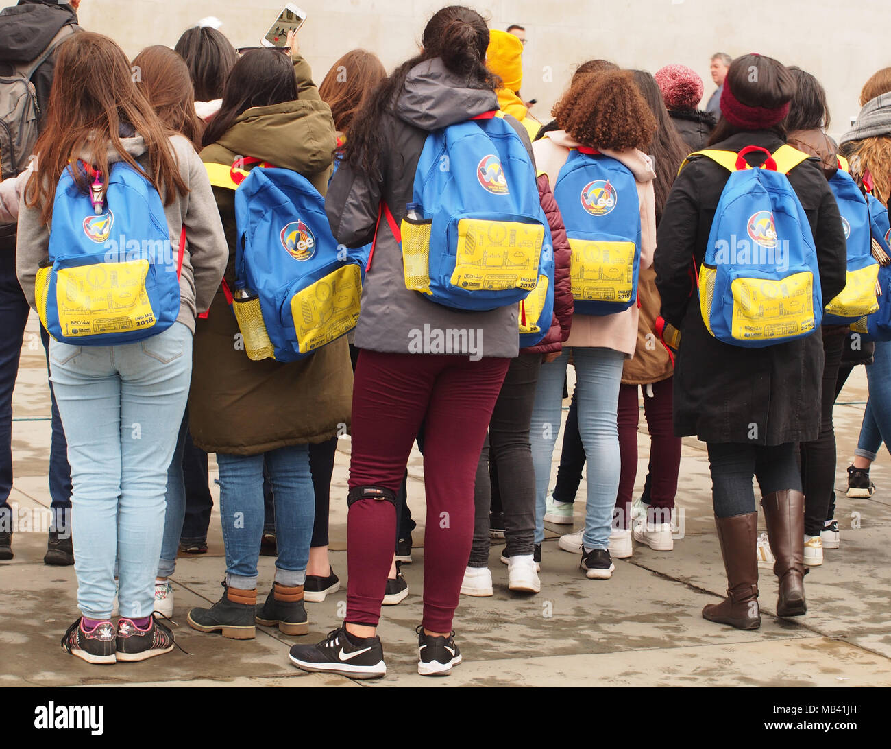 A view of the backs of a group of students visiting London in Trafalgar Square watching one of the entertainers, all carrying identical backpacks Stock Photo