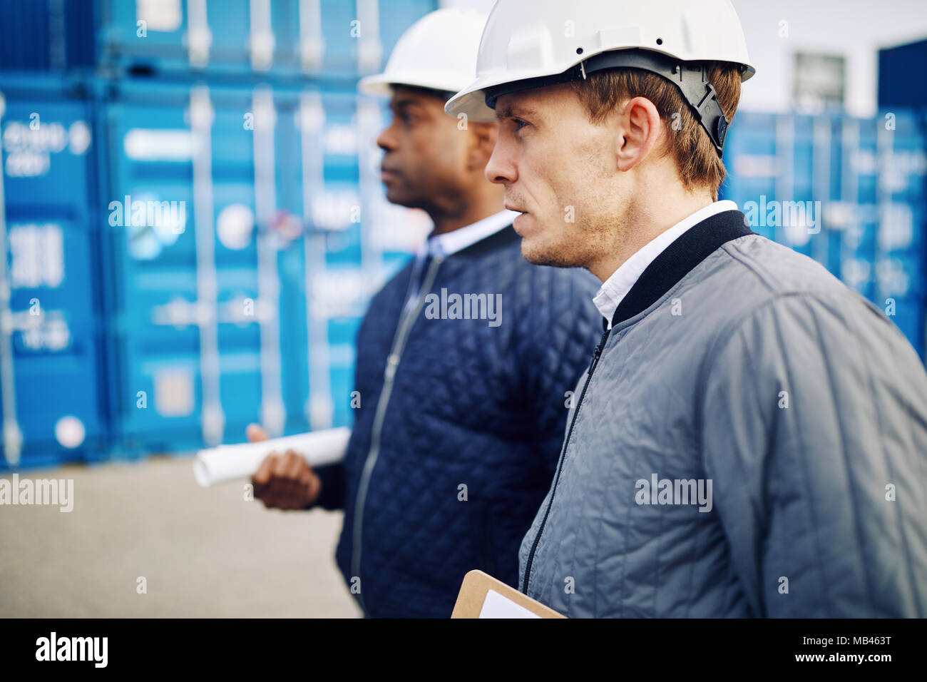 Two engineers wearing hardhats standing together by freight containers on a large commercial shipping dock Stock Photo