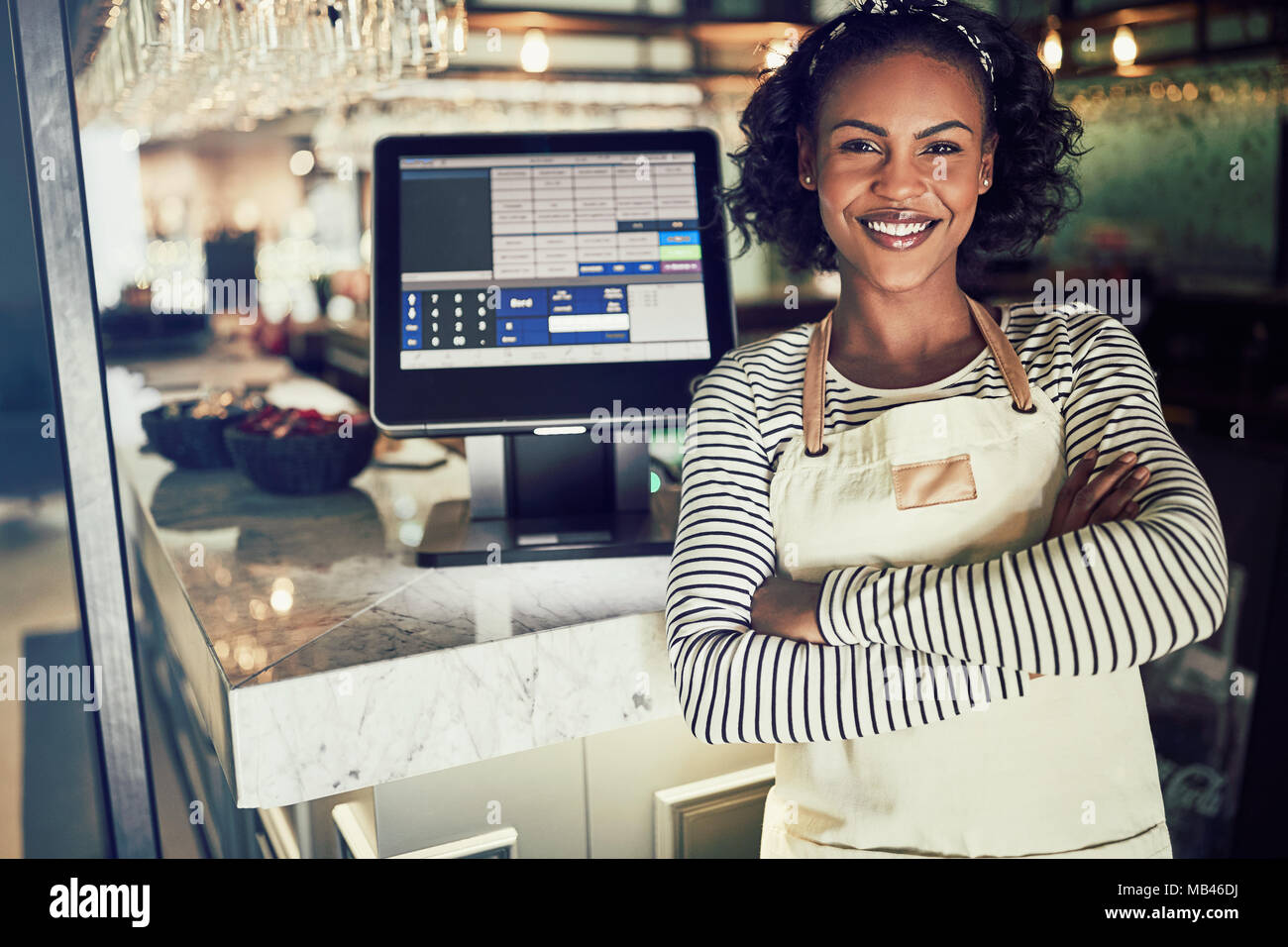 Young African waitress wearing an apron and smiling while standing by a point of sale terminal in a restaurant Stock Photo