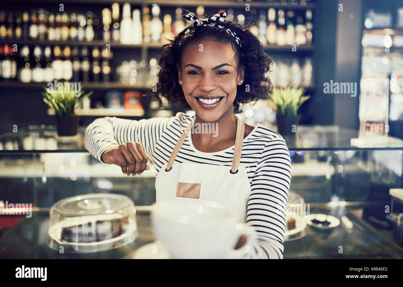 Smiling African barista leaning on the counter of a trendy cafe holding up  a fresh cup of coffee Stock Photo - Alamy