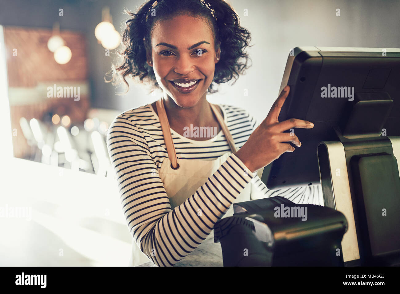 Smiling young African waitress wearing an apron standing by the point of sale terminal of a trendy restaurant Stock Photo