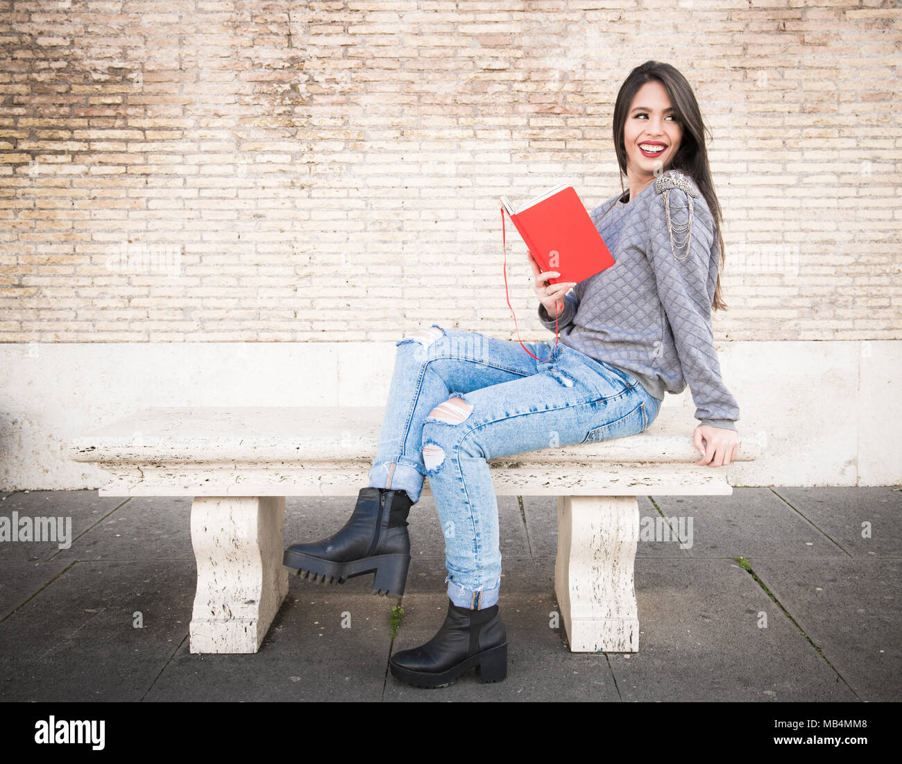 Young latina woman sitting on old marble bench similing and looking out with red travel book in hands Stock Photo